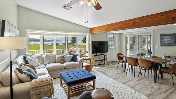 A spacious living room with a large window, a ceiling fan, a TV, and a dining area with wooden furniture. - Honeoye Lake | Hot Tub | Fire Pit (Honeoye)