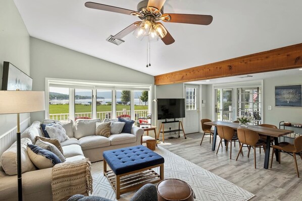 A spacious living room with a large window, a ceiling fan, a TV, and a dining area with wooden furniture.