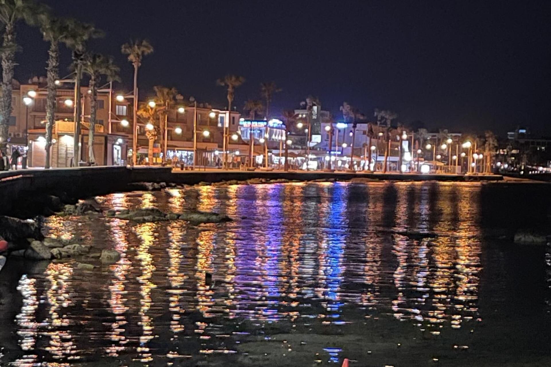 A nighttime waterfront scene with illuminated buildings and a calm body of water reflecting lights.