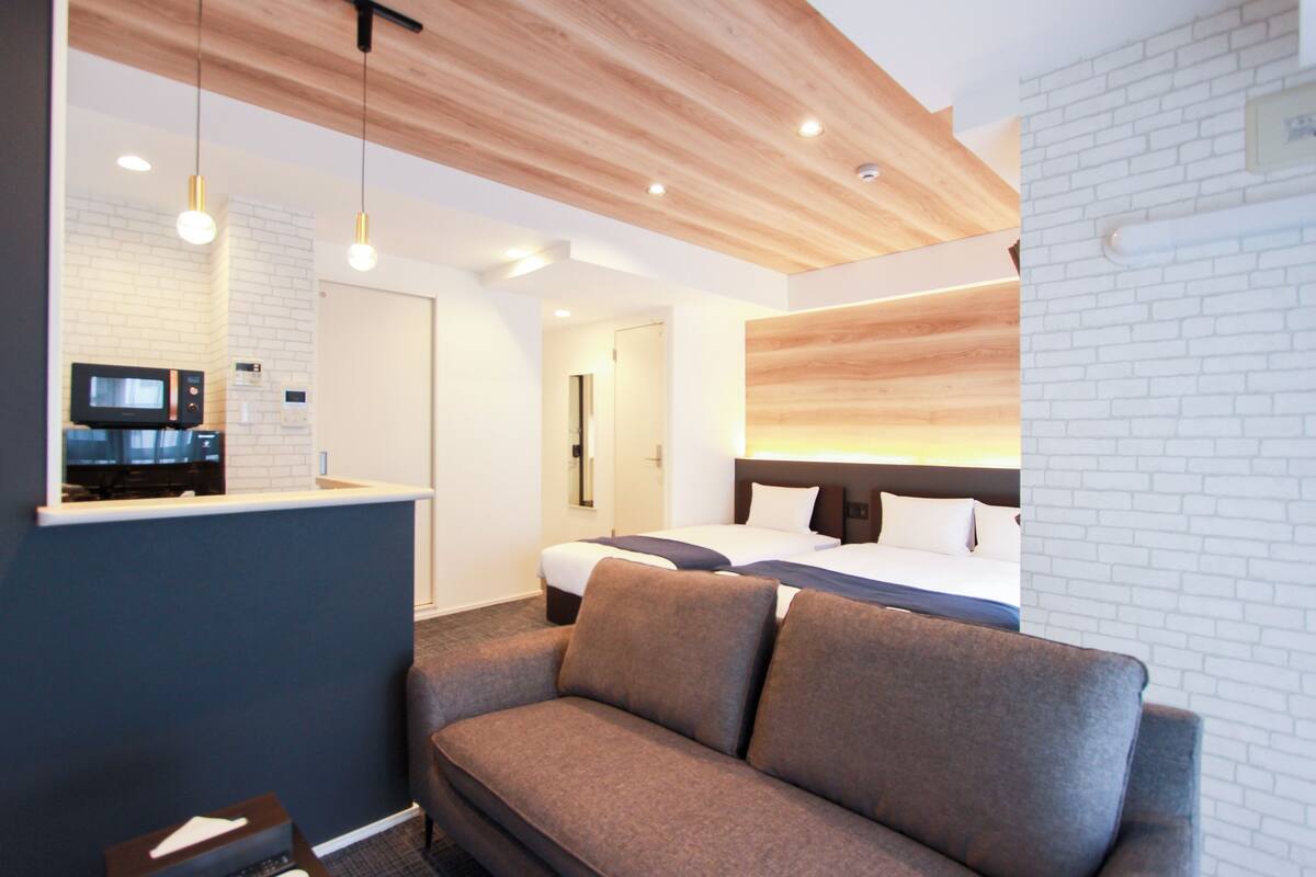 Une chambre d’hôtel moderne avec un plafond en bois, un canapé gris, un lit avec du linge de maison blanc et un téléviseur fixé sur un mur en briques blanches.