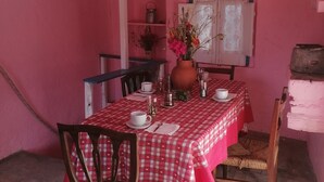 Un comedor con una mesa lista para dos personas, una silla, una ventana con cortinas de encaje y una repisa con una olla de metal.
