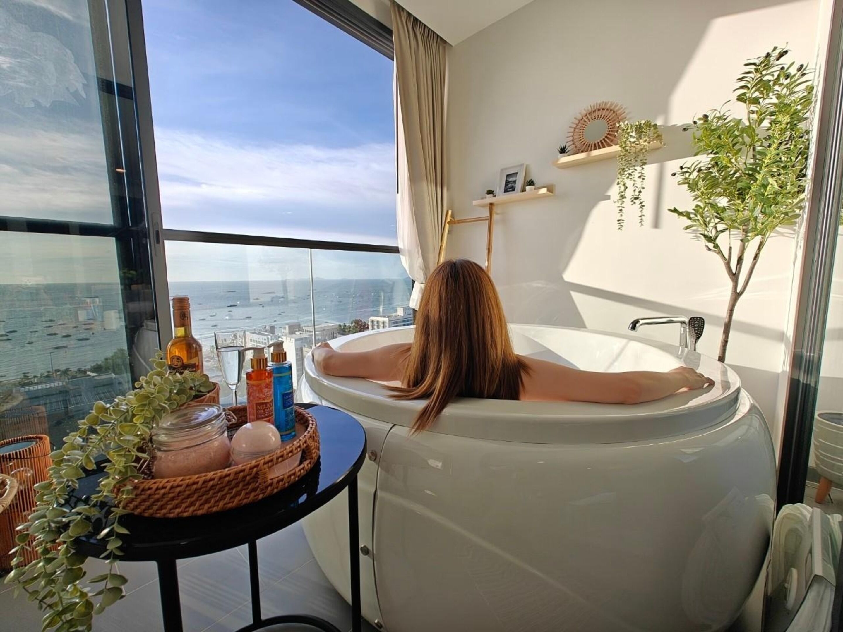 A woman relaxing in a bathtub with a city view, a small table with toiletries, and a plant on the side.