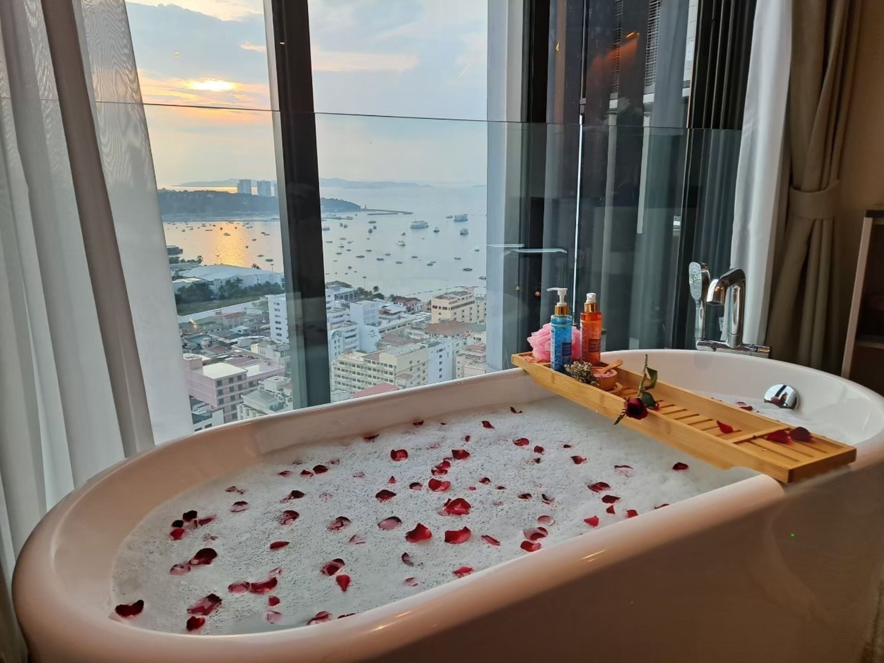 A bathroom with a bathtub filled with water and rose petals, a wooden tray with toiletries, and a view of a cityscape and marina through large windows.