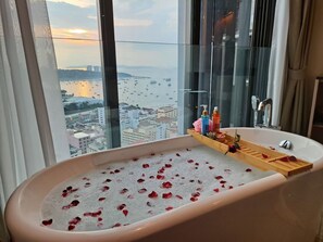 Une salle de bain avec une baignoire remplie d’eau et de pétales de rose, un plateau en bois contenant des articles de toilette, et une vue sur le paysage urbain et le port de plaisance, visible à travers de grandes fenêtres.