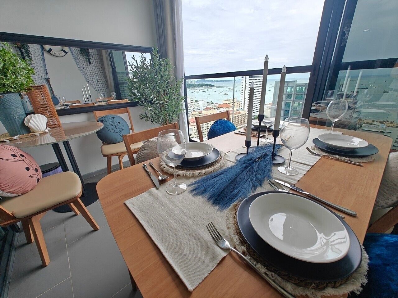 A dining area with a view of the sea, a round table set with plates, glasses, and cutlery, and a chair with a decorative pillow.