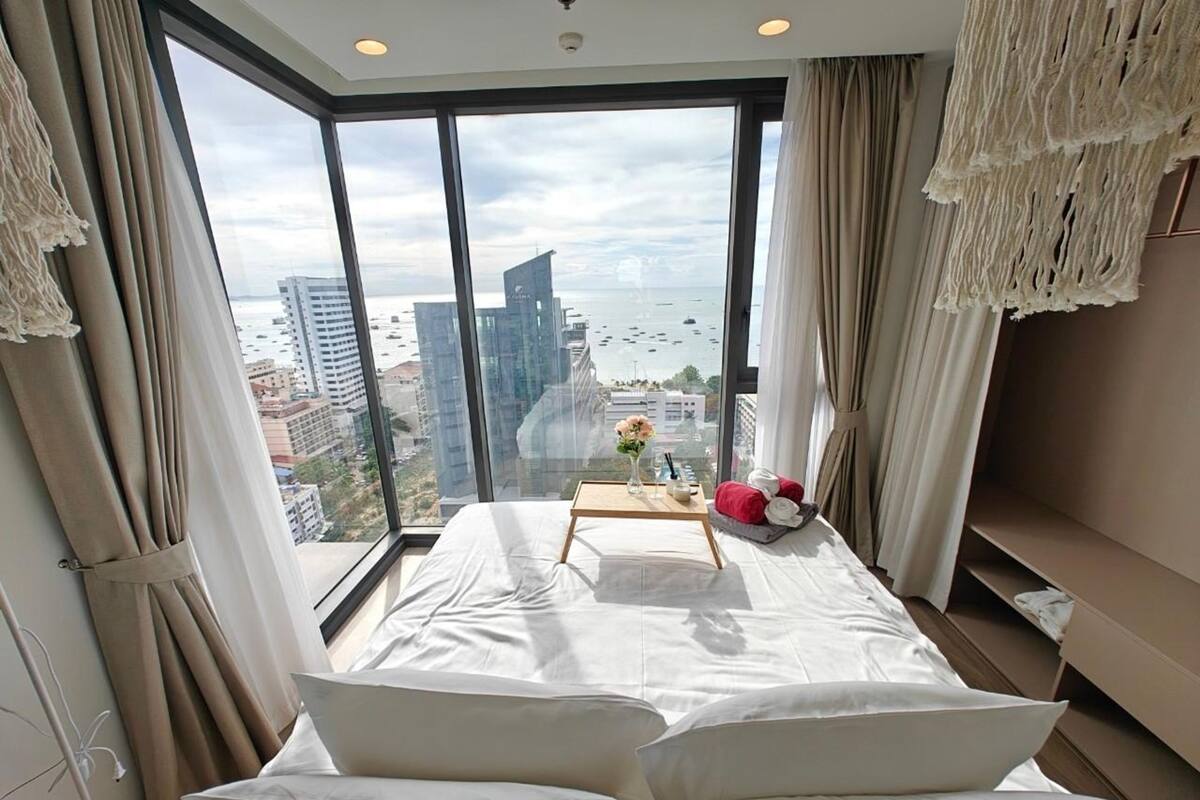 A modern hotel room with a large window offering a city view, a neatly made bed, and a small table with a vase of flowers.