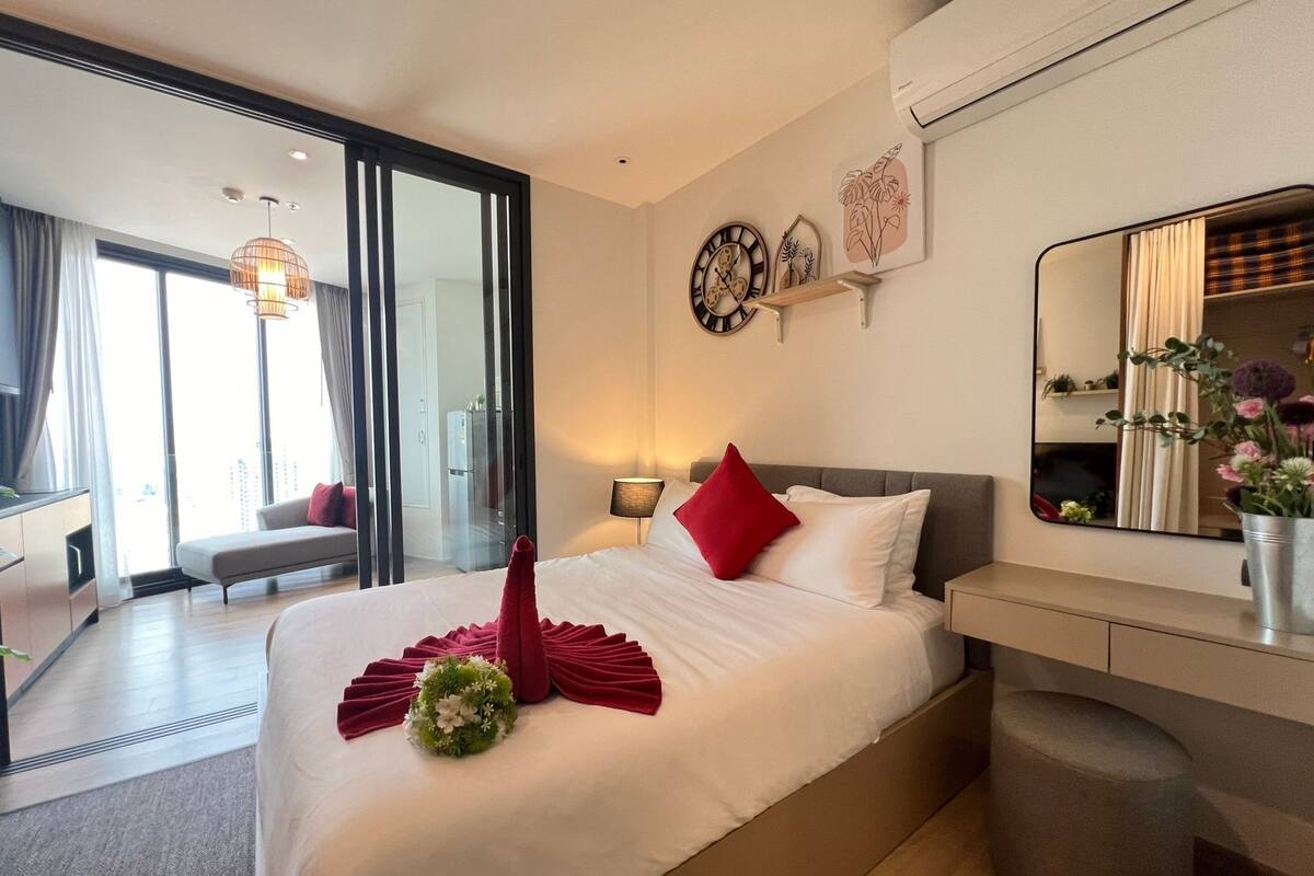 A modern hotel room with a large bed, a bedside table, a mirror, and a view of the city through large windows.