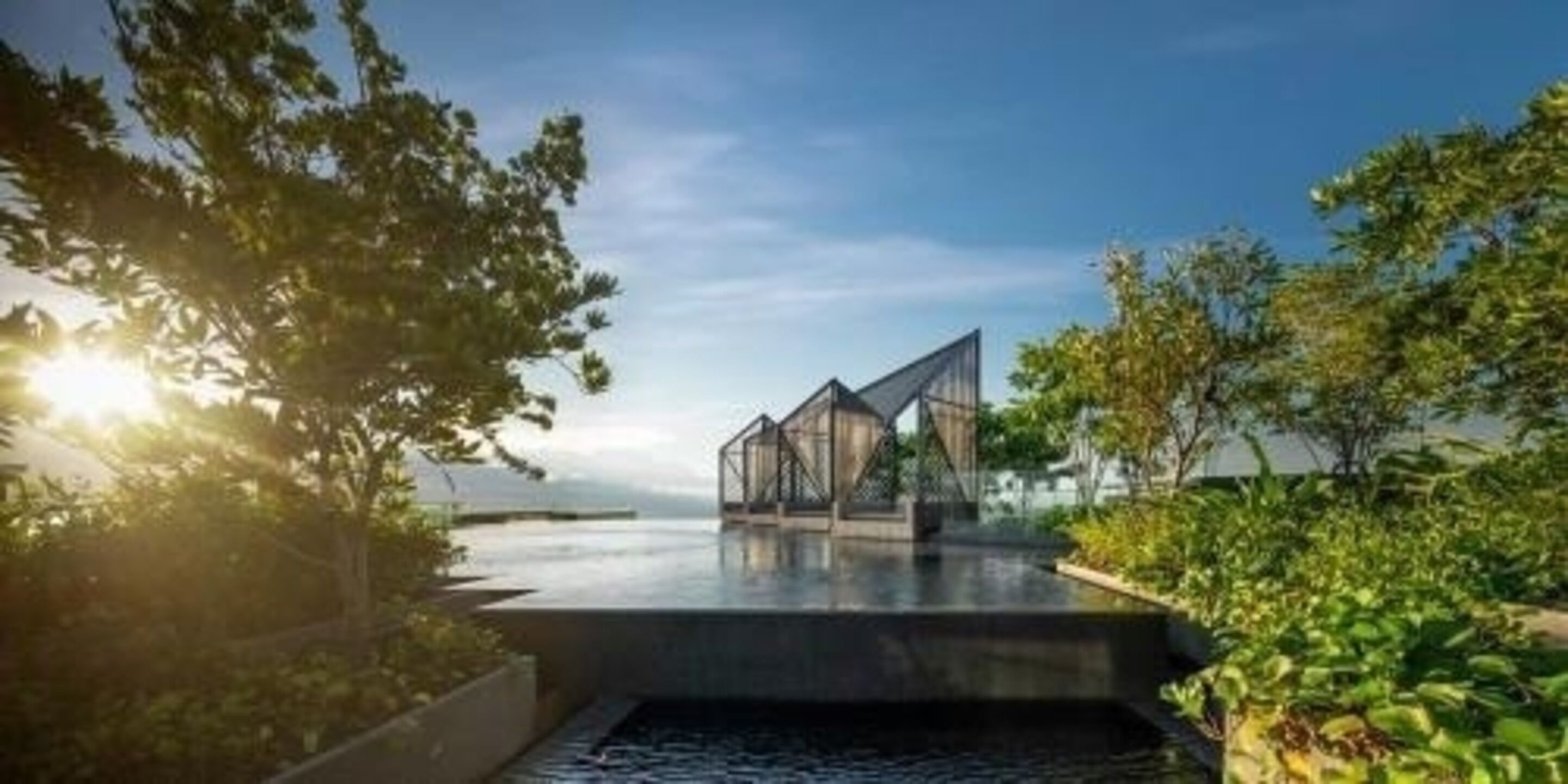A modern architectural structure with a glass facade, situated by a body of water, surrounded by greenery.