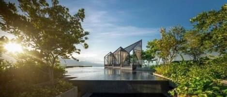 A modern architectural structure with a glass facade, situated by a body of water, surrounded by greenery.