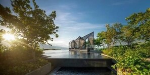 Une structure architecturale moderne dotée d’une façade en verre, située au bord d’un plan d’eau et entourée de verdure.