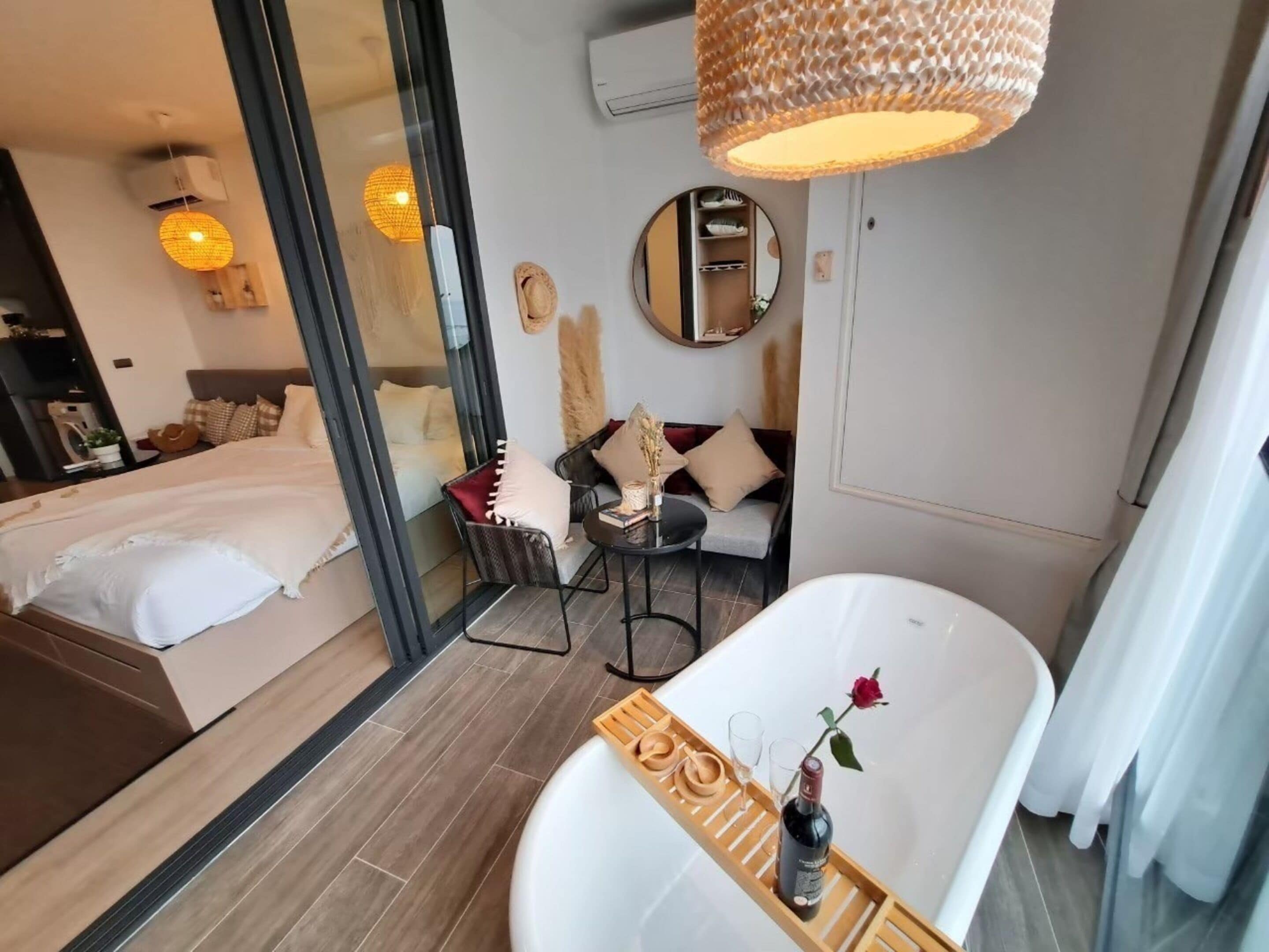 Een moderne hotelkamer met een groot bed, een zithoek met een ronde spiegel, een badkuip met een roos, en een houten dienblad met wijnglazen en een fles.