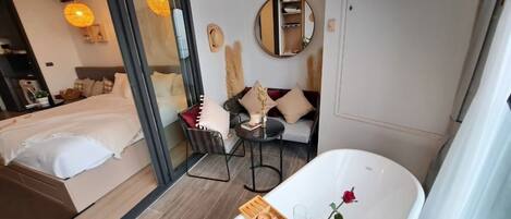 Een moderne hotelkamer met een groot bed, een zithoek met een ronde spiegel, een badkuip met een roos, en een houten dienblad met wijnglazen en een fles.