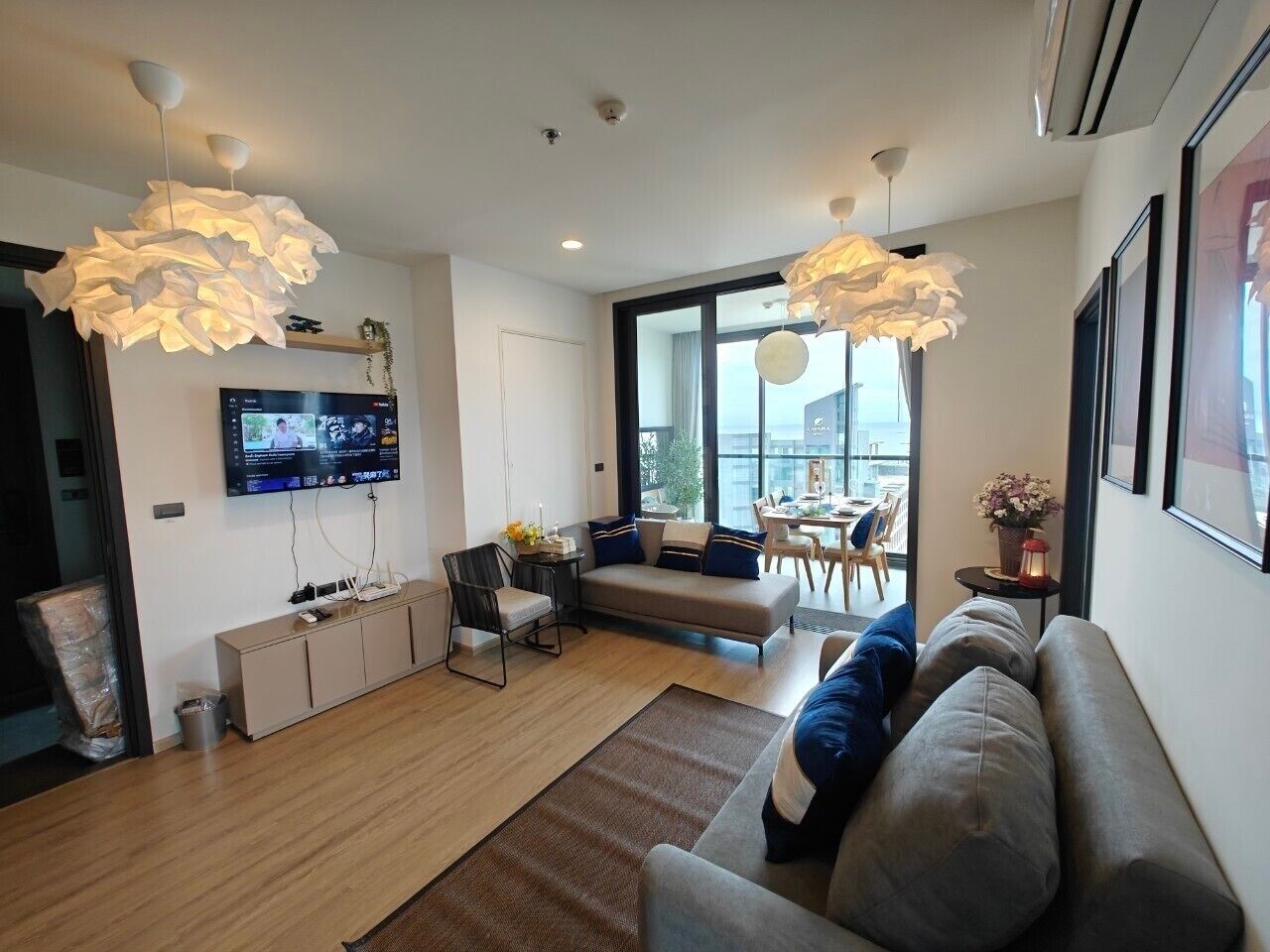 A modern living room with a sofa, a coffee table, and a TV mounted on the wall. There is a dining area with a table and chairs visible through a glass door.