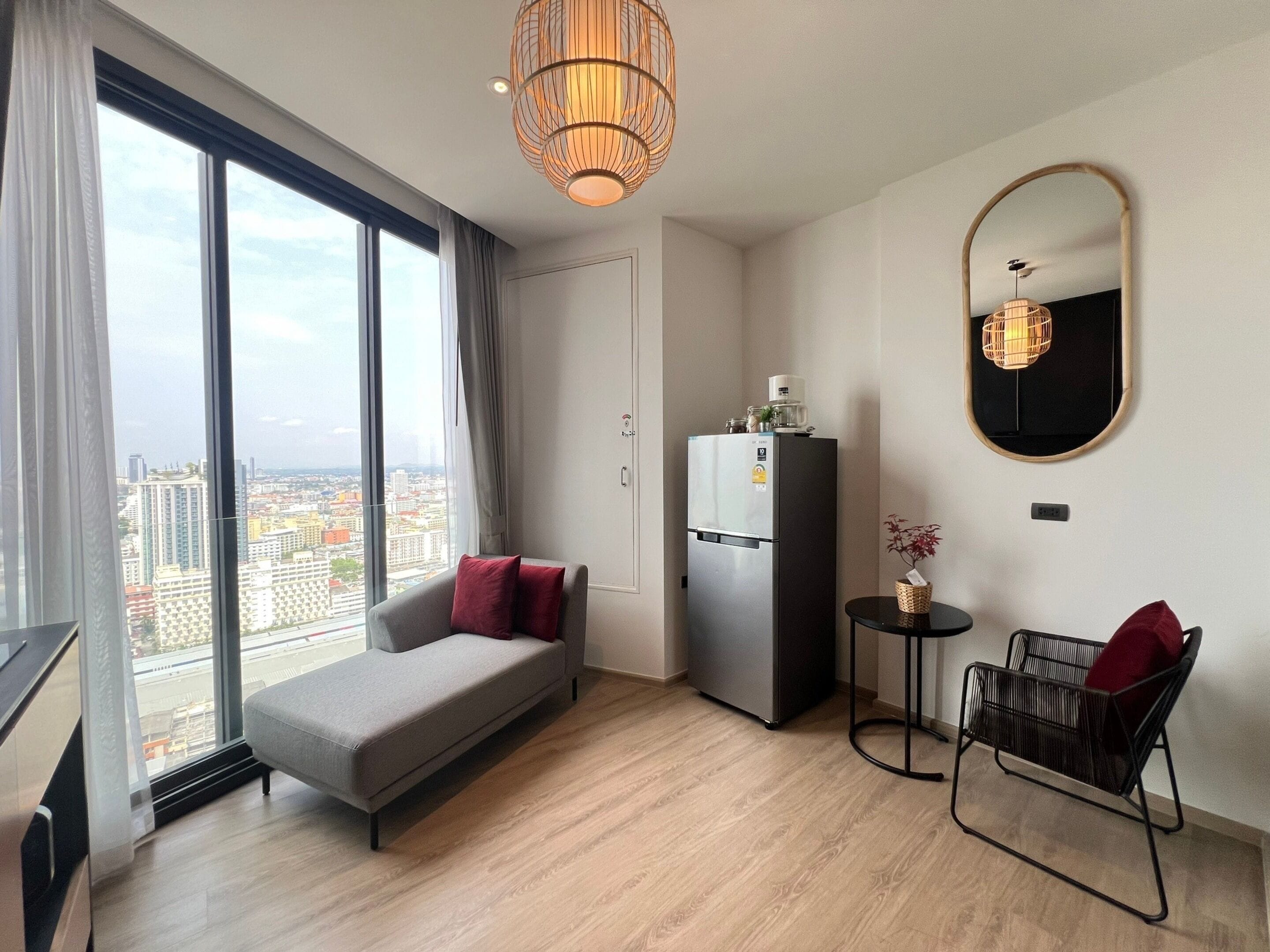 A modern living room with a sofa, a small table, a chair, a refrigerator, and a large window offering a city view.