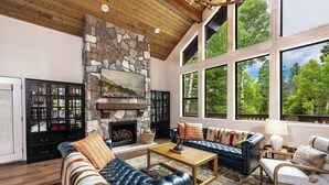 Amplia sala de estar con chimenea de piedra, sofás de cuero y ventanas grandes con vistas al bosque.