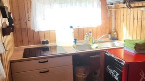 A compact kitchen with wooden walls, a window with lace curtains, a sink, a stove, and a red Coca-Cola refrigerator.