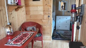 A cozy wooden cabin interior with a dining area, a red wicker chair, a small kitchen with a stove, and a decorative rooster wall hanging.