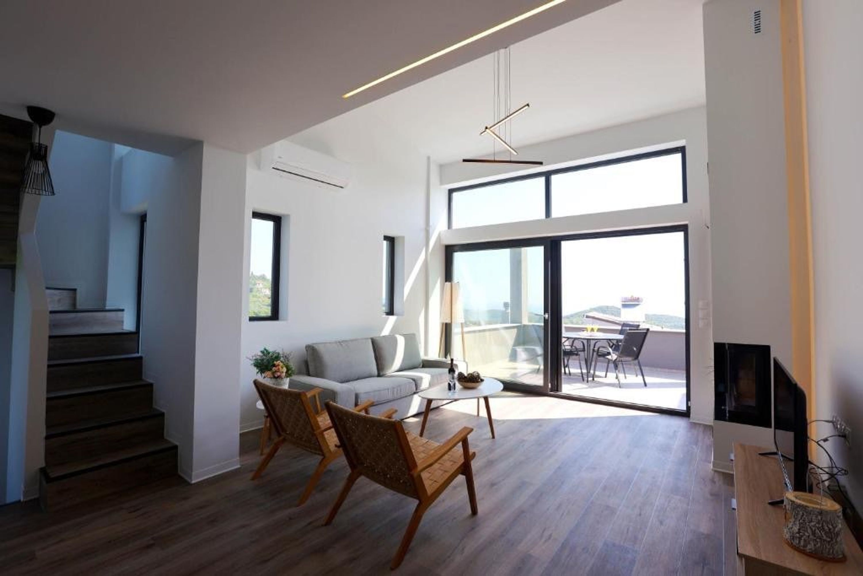 A spacious living area with a sofa, two chairs, a coffee table, and a TV on a wooden stand. There is a staircase leading to another level and a balcony with a table and chairs.