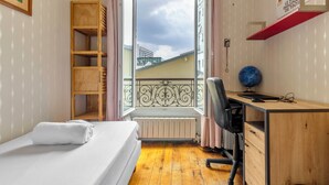 Quarto com cama, escrivaninha, cadeira, radiador, janela com vista para edifícios e prateleira com um globo.