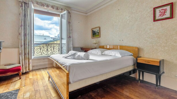 Quarto com cama de madeira, mesa de cabeceira, um banquinho vermelho, janela com cortinas e um quadro na parede.