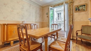 Uma sala de jantar com mesa e cadeiras de madeira, um candelabro e vista para um edifício através de uma porta aberta.