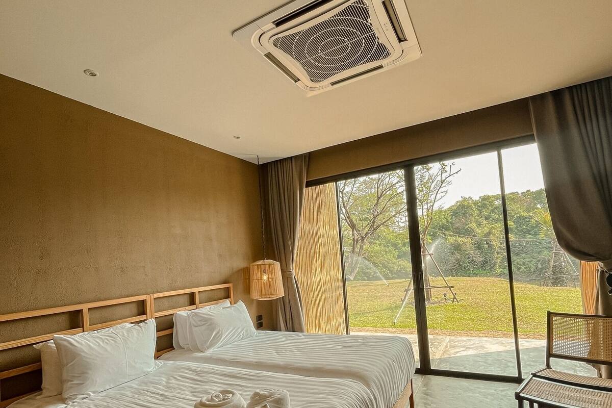 A hotel room with a large bed, a wooden headboard, a bedside table, a lamp, and a view of the outdoors through a large window.