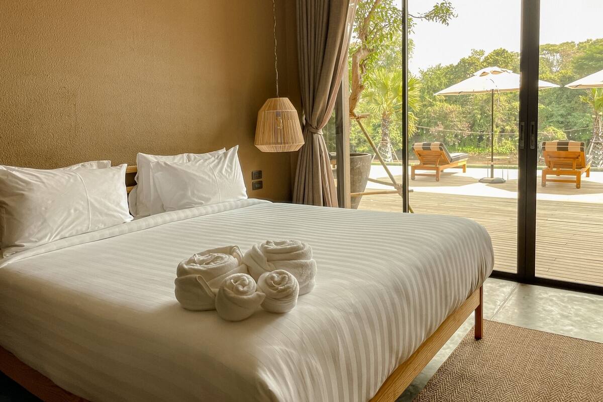 A hotel room with a large bed, white linens, and a view of outdoor seating and greenery.