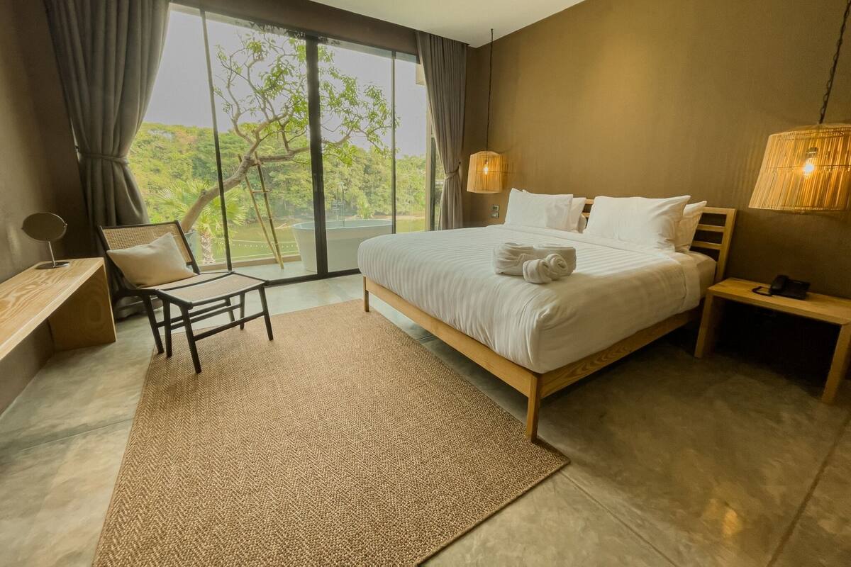 A modern bedroom with a large bed, a desk, a chair, and a view of greenery through the window.