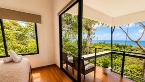 Une chambre avec une grande fenêtre, un balcon offrant une vue sur la verdure et la mer, et un lit avec une serviette enroulée.