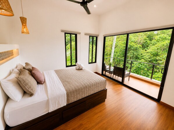 Une chambre avec un grand lit, deux fenêtres, un balcon et un ventilateur de plafond.