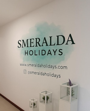 Un muro con la scritta 'SMERALDA HOLIDAYS' e un indirizzo web. Sono presenti tre vetrine espositive con piante.