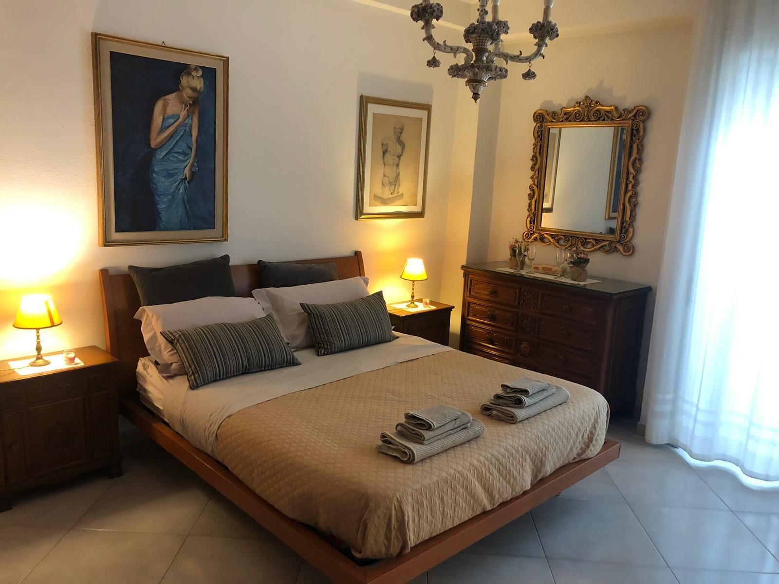 A well-lit bedroom with a large bed, two bedside lamps, a dresser, a mirror, and two framed artworks on the wall.