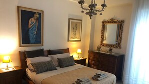 Une chambre bien éclairée, avec un grand lit, deux lampes de chevet, une commode, un miroir et deux œuvres d’art encadrées accrochées au mur.
