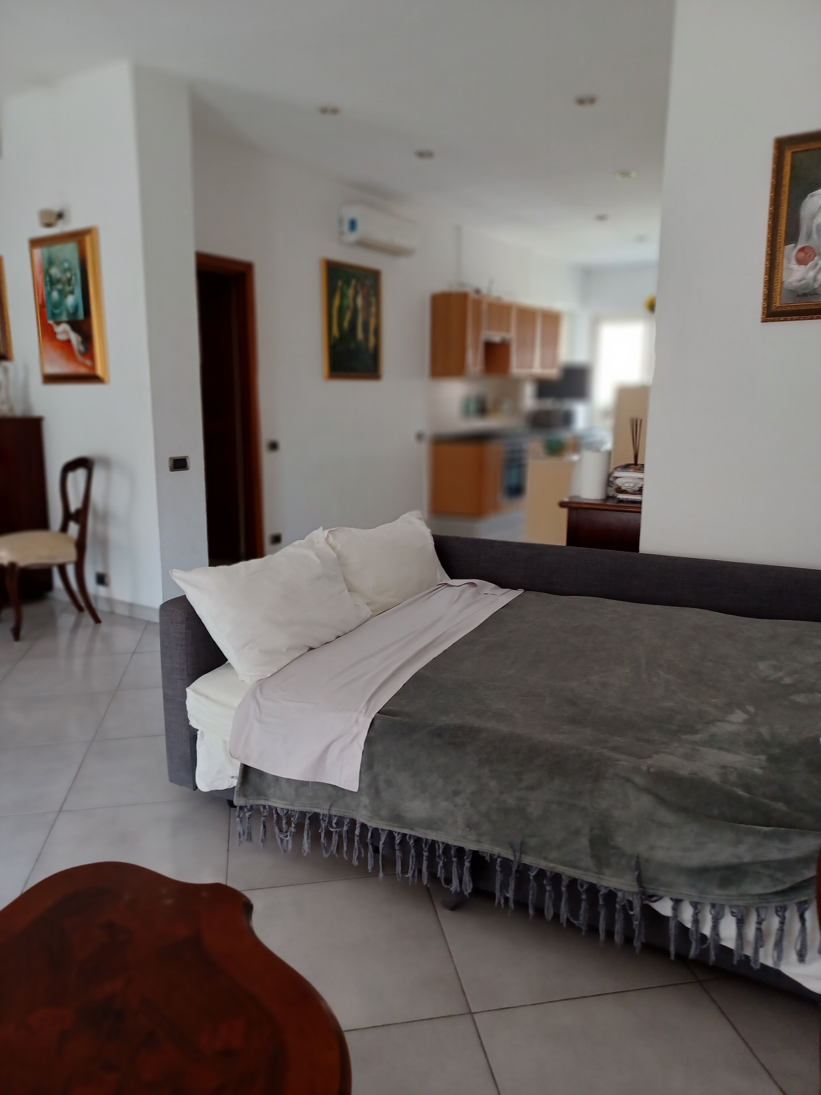 A modern bedroom with a grey sofa bed, a wooden chair, and a dining table in the background.