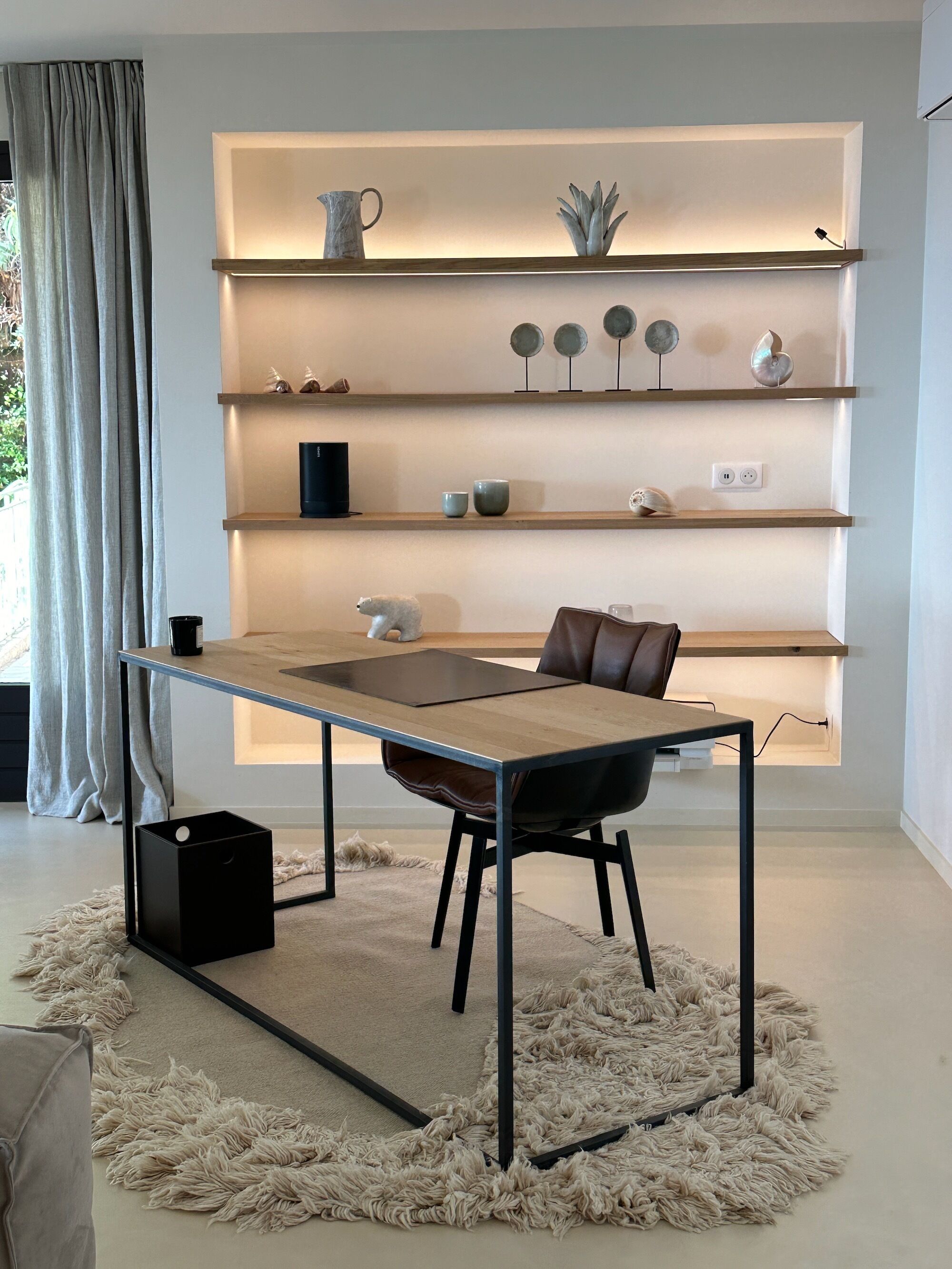 Una sala de estar moderna con una mesa de madera, un sillón de cuero y un alfombrado mullido. Se observan estantes con objetos decorativos y una ventana con cortinas.