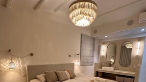 Une chambre à coucher avec un grand lit, des tables de chevet, un lustre suspendu, une coiffeuse avec un miroir et une fenêtre avec des rideaux.
