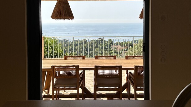 Une pièce avec vue sur la mer, visible par une fenêtre, une table en bois avec des chaises et un luminaire suspendu.