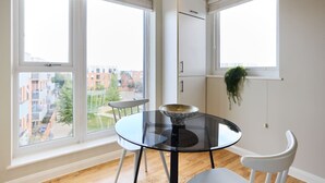 A modern dining area with a glass table, white chairs, and a view of buildings through large windows. - The Enfield Sanctuary - Modern 1bdr Flat With Balcony (Enfield)