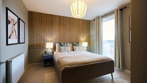 A hotel room with a large bed, a bedside table with lamps, a wooden panel wall, and a view of the city through the window. - The Enfield Sanctuary - Modern 1bdr Flat With Balcony (Enfield)