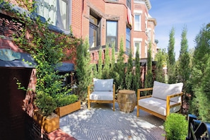A cozy outdoor seating area with two wooden chairs and a round table, surrounded by potted plants and a patterned rug.