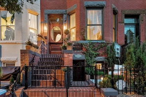 A two-story brick building with a wooden front door, a small porch with steps, and a black metal gate.