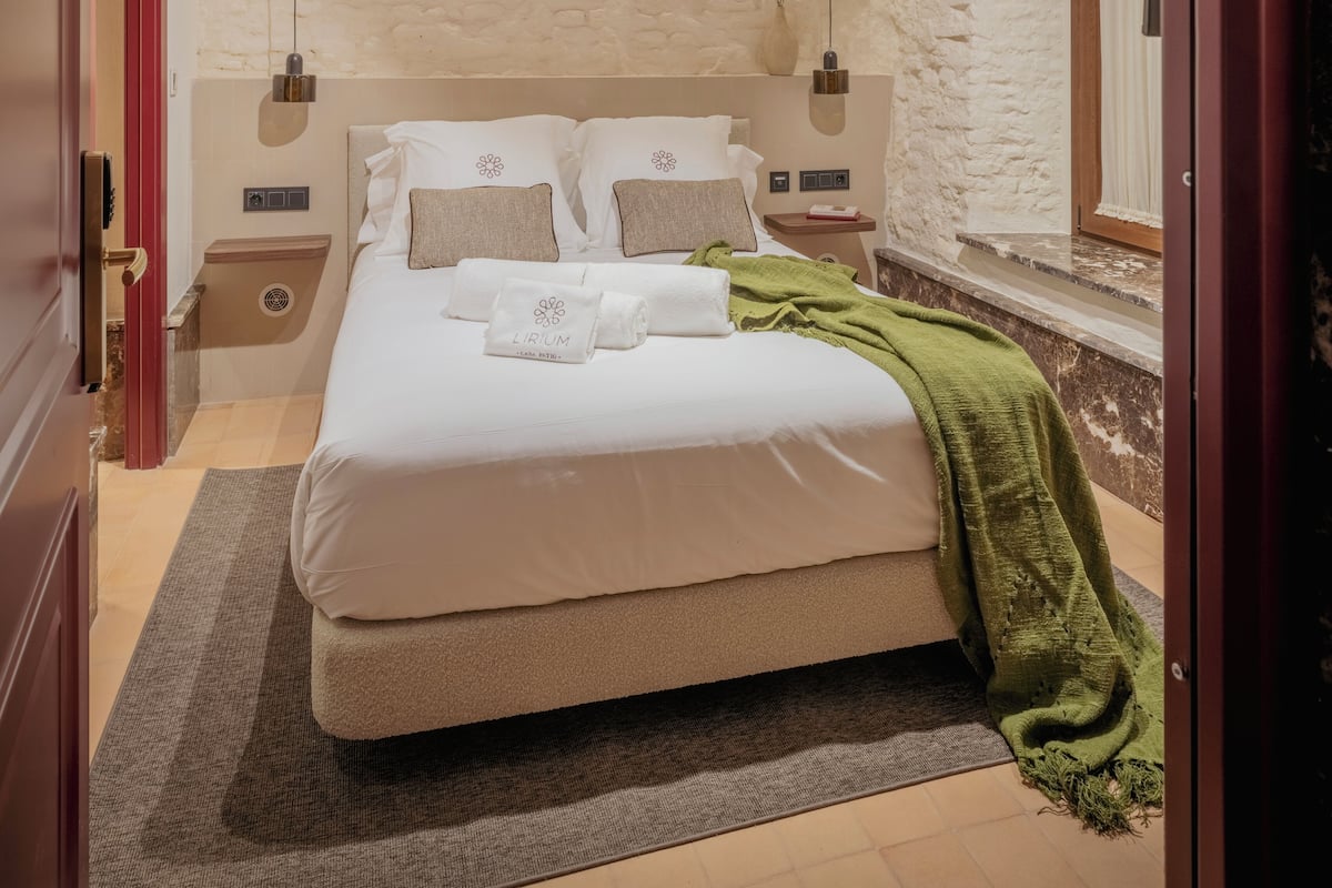 A neatly made bed with a green throw blanket, a white headboard, and a stone wall behind it.