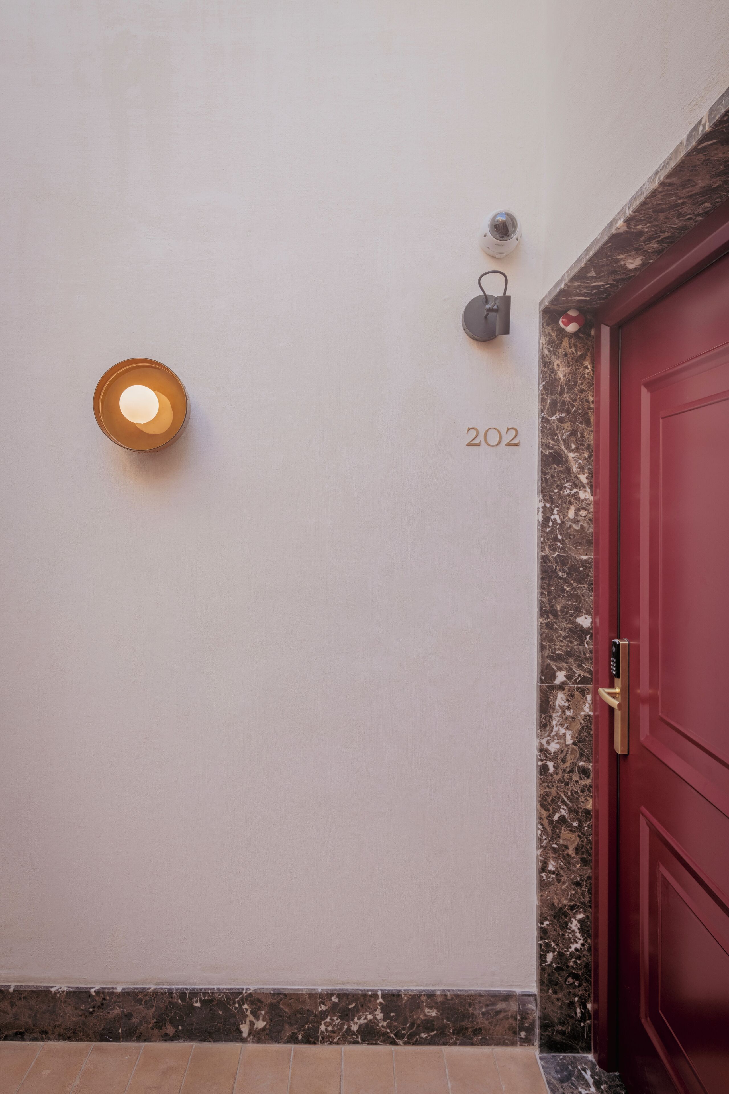 Un couloir avec une porte rouge, le numéro « 202 » peint sur le mur, une caméra de surveillance et un luminaire.