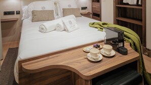 Un lit bien fait, avec du linge de lit blanc et une serviette verte, une table de chevet en bois sur laquelle se trouvent une tasse et une soucoupe, et une robe suspendue.
