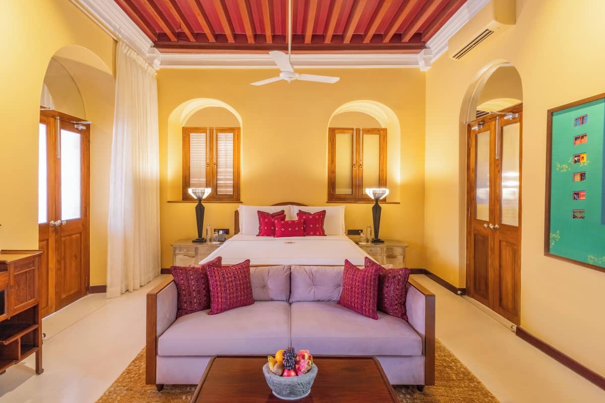 Une chambre d’hôtel avec un lit, un canapé, une table basse avec des fruits et deux lampes.