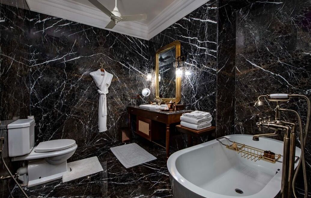 Un baño con pared de mármol, inodoro blanco, lavamanos con grifo dorado y espejo con iluminación.