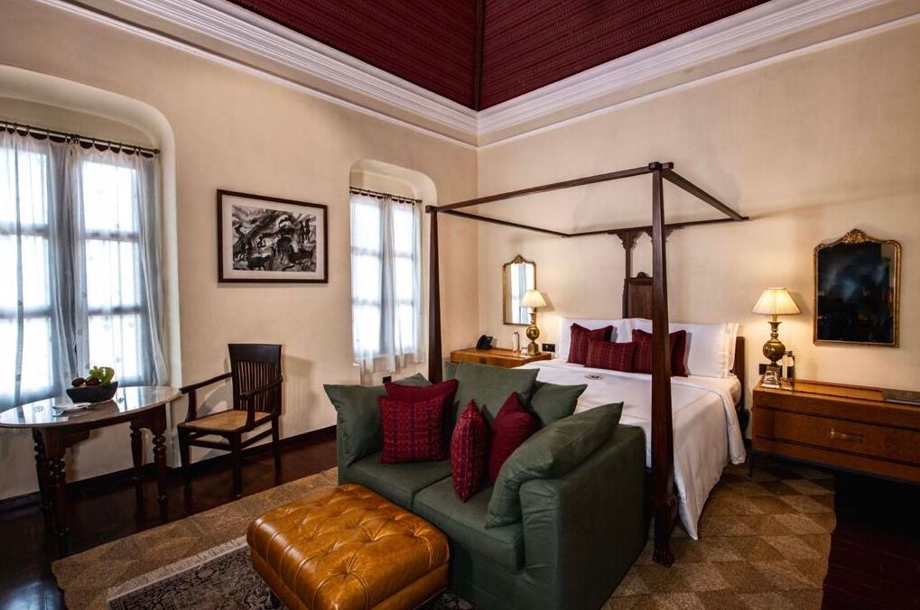 A four-poster bed with a canopy, a green sofa with red cushions, a wooden chair, a small table with a bowl of fruit, and a lamp on a nightstand.