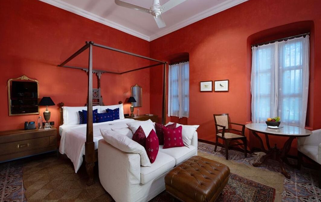 A room with a four-poster bed, a white sofa, a brown ottoman, a round table with a fruit bowl, and a chair.