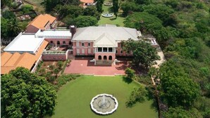 Un grand bâtiment doté d’une cour intérieure et d’une fontaine, entouré d’une végétation luxuriante et avec une étendue d’eau en arrière-plan.