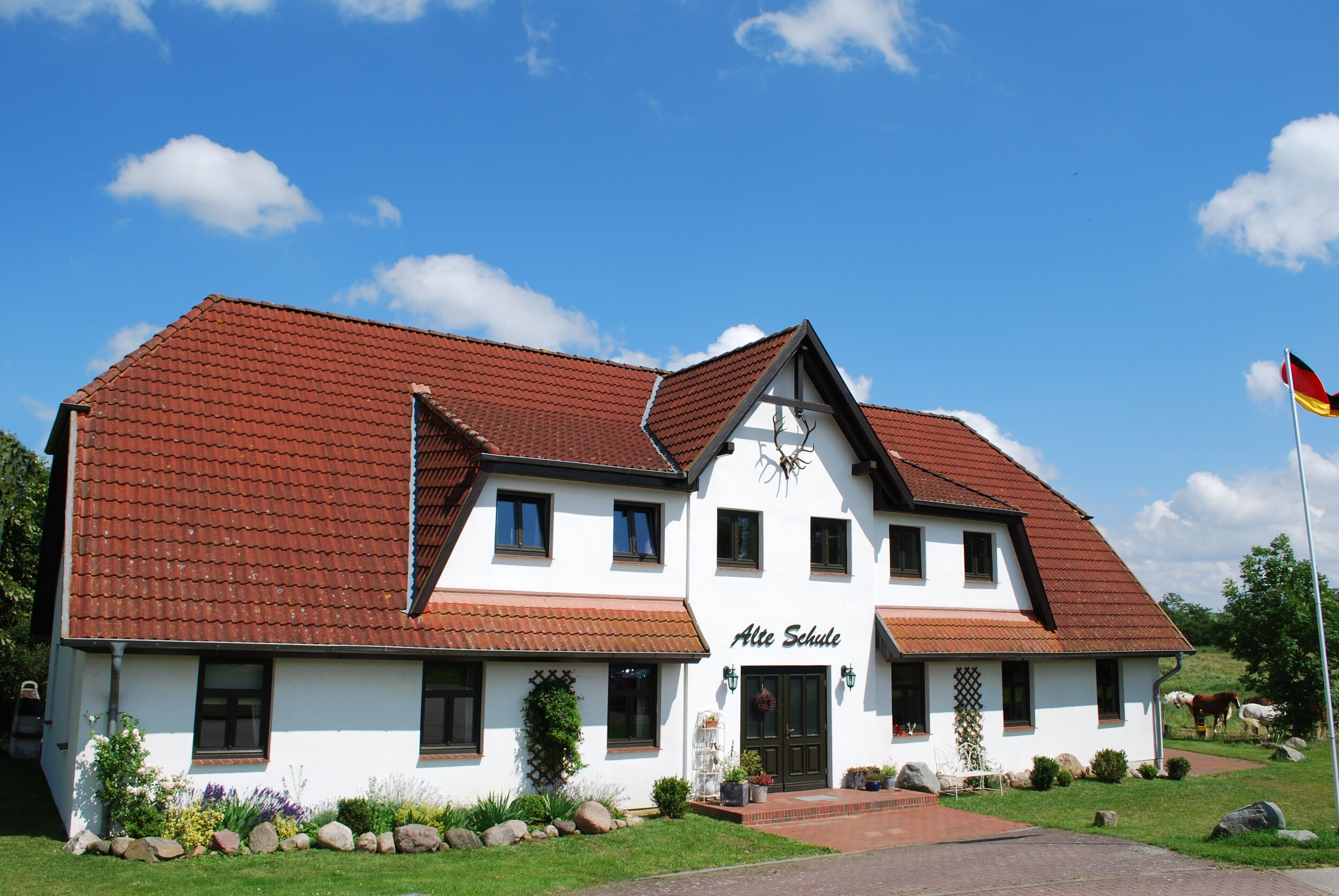 Un bâtiment à deux étages, avec un toit recouvert de tuiles rouges et une enseigne indiquant « Alte Schule ».